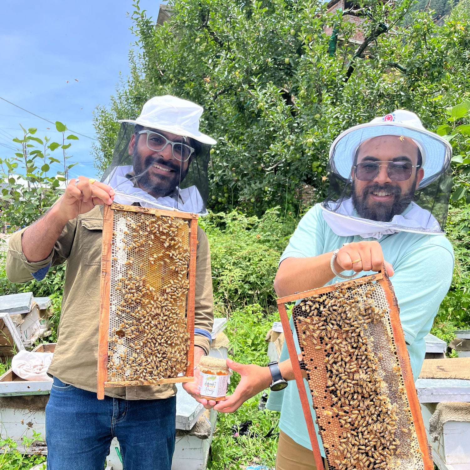 Two beekeepers holding honeycomb frames outdoors with greenery in the background